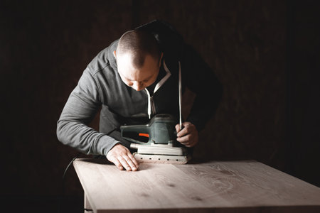 A carpenter works in a workshop. Joiner's grinders, furniture manufacturing. A carpenter is grinding a wooden part with an electric sander.の写真素材