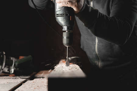 A man works with a drill in his workshop. Carpenter drills with a hand power tool in a dark room with directional lightの写真素材