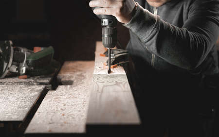 A man works with a drill in his workshop. Carpenter drills with a hand power tool in a dark room with directional lightの写真素材