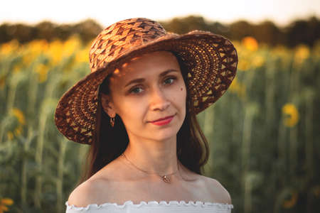 A young girl in a white dress and hat in a field of sunflowers at sunset. Portrait of a woman with a slim figure on a background of yellow flowersの写真素材