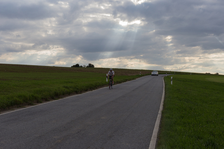 on a very sunny day in august in south germany you see cyclist and car on a road  beside green fields with contrastful cloudy skyの写真素材