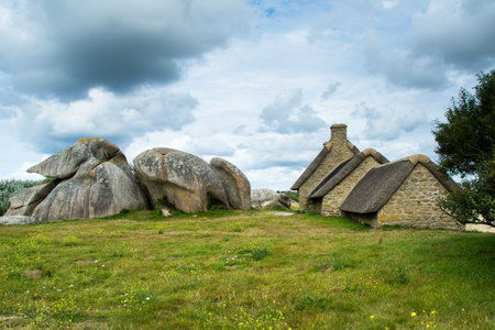 Old stone house with thatched roof and green meadow under cloudy sky in Menehan, Bretagne, Franceの写真素材