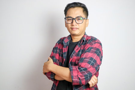 A young Asian man, wearing a casual shirt, smiles confidently with his arms crossed. The background...