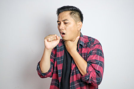 Young Asian man in casual t-shirt coughing into his fist while holding his chest, indicating...