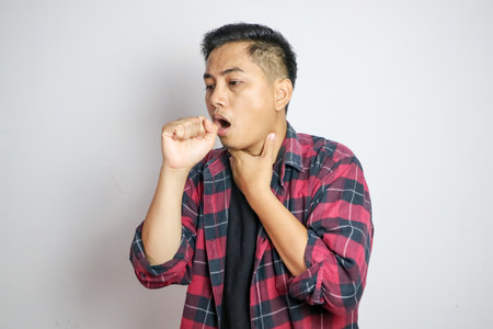 Young Asian man in casual outfit coughing into his fist while holding his chest, indicating discomfort or illness. Studio shot on plain backgroundの写真素材
