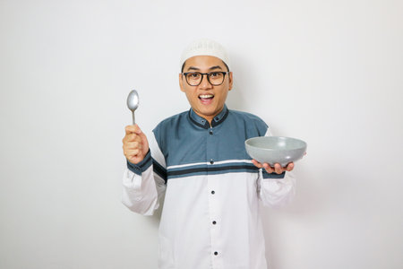 Excited young Asian Muslim man wearing Arabic clothes standing while holding bowl and saying yes it's time to break fast isolated on white background. People religious Islamic lifestyle conceptの写真素材
