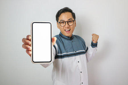 Smiling young Asian Muslim man pointing finger at display mobile phone screen isolated on white...
