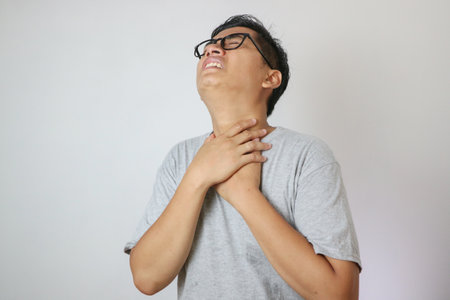 Portrait of young asian man in casual white t-shirt isolated on white background. He had a sore throat and touched his throat. Difficult to swallowの写真素材