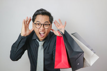 Portrait of young Asian man shouting and holding shopping bags in pose on white backgroundの写真素材