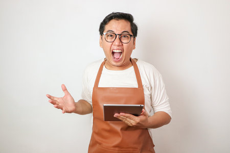 Asian waiter man in apron holding tablet yelling with aggressive expression and arms raised.の写真素材