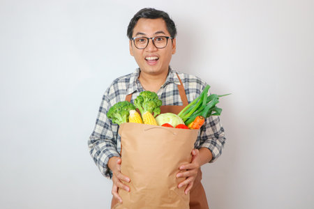 Young smiling happy asian man wear shirt and apron hold brown craft bag for takeaway mock up with food products show thumb up gesture isolated on plain blue background. Delivery service from shop or restaurantの写真素材