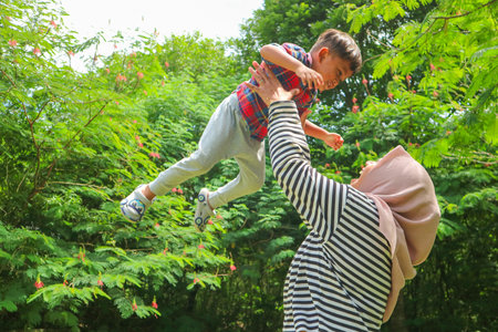 A mother woman plays with a child in nature, lifting and tossing him in her arms. Toddler baby and mom play on the green grass in the summer park. Kid aged about two years (one year eleven months)の写真素材