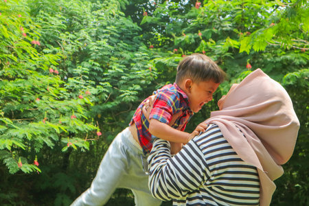 A mother woman plays with a child in nature, lifting and tossing him in her arms. Toddler baby and mom play on the green grass in the summer park. Kid aged about two years (one year eleven months)の写真素材