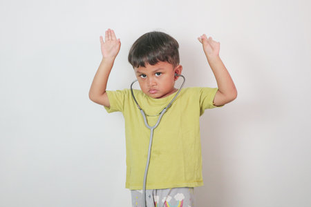 Smiling little boy in medical uniform playing with stethoscope and hands up isolated on white