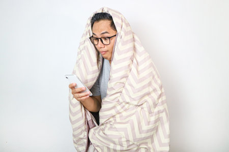 Portrait of young sleepy tired man under the blanket using smartphone at late night, can not sleep/ Insomnia, nomophobia, sleep disorder concept/ Dependency on a cell phoneの写真素材