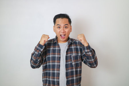 Excited Asian man clenching his fists and screaming in joy, showing a gesture of celebrating victory...