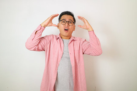 Mind blowing. Wow and shocked face expression of young Asian Malay man with both hands up and opened mouth point isolated white background. Advertising model concept.の写真素材