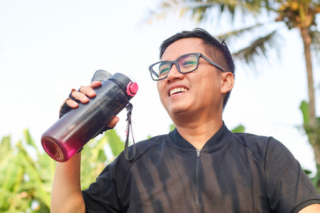 Asian man in sportswear drinking water from a bottle during jogging exercise at public park in the morning. Healthy guy athlete enjoy outdoor lifestyle sport training workout running in the city.の写真素材