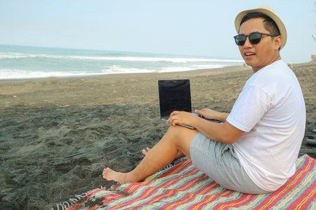 Lifestyle young asian man blogger working and using keyboard on laptop while sitting on the beautiful beach, freelance travel on holiday summer, blue sky outdoors. Summer Vacation Concept.の写真素材