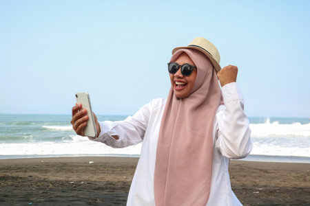 Excited beautiful Asian woman in green sweater and hijab using a mobile phone, celebrating success, getting good news beach backgroundの写真素材