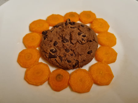Delicious chocolate chip cookie surrounded by thin carrot slices on a white plateの写真素材