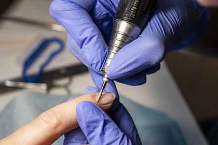 Nail service. Electric nail file drill used in beauty studio. Master of manicure processing operation in blue gloves, debris flying around. Close-up image.の写真素材