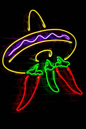 Glowing Neon stylized mexican sombrero and red peppers and blurred lights on black brick wall background. Dark tones vintage image.の写真素材