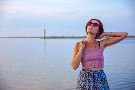 A beautiful young girl in a pink T-shirt and a blue skirt poses on the embankmentの写真素材