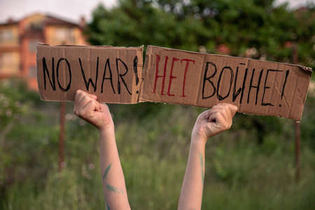A boy with a grimy face and sad eyes holds a cardboard poster with the inscription NO WAR in English and russian in his hands. A small boy of European appearance opposes military action.の写真素材