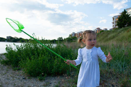 A beautiful little girl in a white dress and with a green scoop-net in her hands on the beach and against the background of green grass and blue sky. Beautiful baby-girl with net.の写真素材