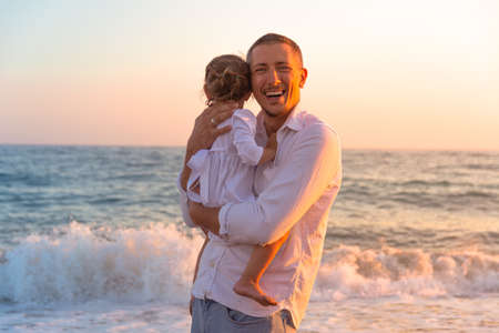 A young dad with a little daughter walks along the sea beach in the sunset rays. A father and a two-year-old daughter in white clothes are walking along the surf line against the backdrop of sea waves. Photo about family values, father's day.の写真素材