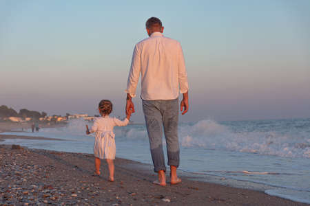 A young dad with a little daughter walks along the sea beach in the sunset rays. A father and a two-year-old daughter in white clothes are walking along the surf line against the backdrop of sea waves. Photo about family values, father's day.の写真素材