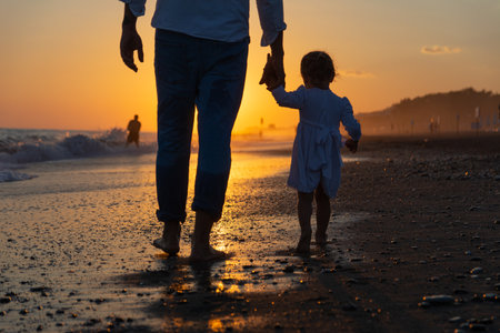 A young dad with a little daughter walks along the sea beach in the sunset rays. A father and a two-year-old daughter in white clothes are walking along the surf line against the backdrop of sea waves. Photo about family values, father's day.の写真素材