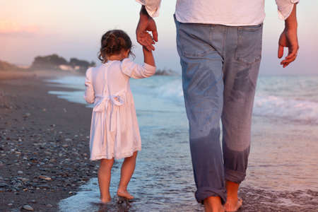 A young dad with a little daughter walks along the sea beach in the sunset rays. A father and a two-year-old daughter in white clothes are walking along the surf line against the backdrop of sea waves. Photo about family values, father's day.の写真素材