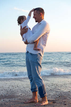 A young dad with a little daughter walks along the sea beach in the sunset rays. A father and a two-year-old daughter in white clothes are walking along the surf line against the backdrop of sea waves. Photo about family values, father's day.の写真素材