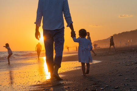 A young dad with a little daughter walks along the sea beach in the sunset rays. A father and a two-year-old daughter in white clothes are walking along the surf line against the backdrop of sea waves. Photo about family values, father's day.の写真素材