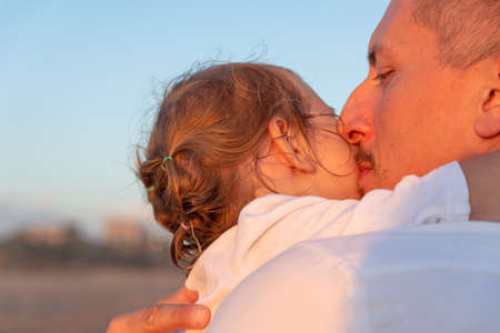 A young dad with a little daughter walks along the sea beach in the sunset rays. A father and a two-year-old daughter in white clothes are walking along the surf line against the backdrop of sea waves. Photo about family values, father's day.の写真素材