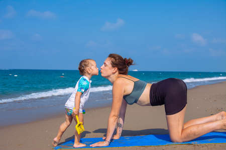 A beautiful girl is doing yoga on the beach near the sea. A girl in tracksuit does yoga and kissing her daughter exercises on the sand, and against the backdrop of the sea surf and a beautiful sky.の写真素材