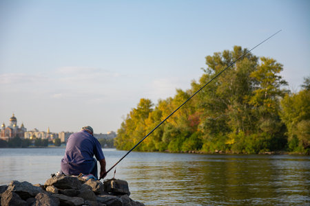 The fisherman sits on the river on the pier relaxes and enjoys fishing. Back view of fisherman with a lot of fishing rods and a net for fish. Fishing on the river.の写真素材