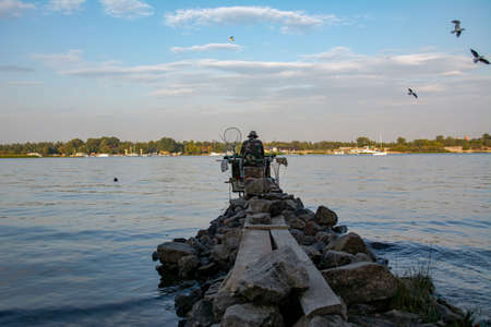 The fisherman sits on the river on the pier relaxes and enjoys fishing. Back view of fisherman with a lot of fishing rods and a net for fish. Fishing on the river.の写真素材