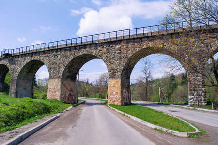 Railway Bridge-Viaduct in Carpathian village Vorokhta. The road under stone bridge, green grass and trees at sunny spring day.の写真素材