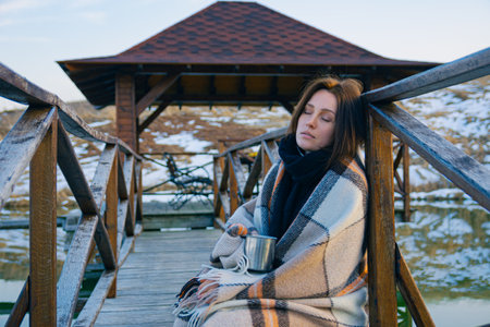 A beautiful thoughtful and sad girl wrapped in a plaid walks along the bridge over the pond with a metal mug of tea on a cold autumn day. Against the background of the forest and nature of Ukraine.の写真素材