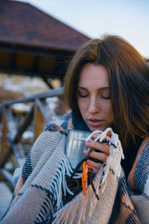 A beautiful thoughtful and sad girl wrapped in a plaid walks along the bridge over the pond with a...