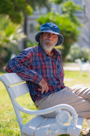 An old man with a beard in a panama hat and a plaid shirt sits on a park bench on a summer sunny day.の写真素材