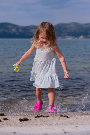 Little cute girl in dress and pink sandals on the sea beach. Beautiful Caucasian girl jumps in shallow water in sea water and splashes of water fly to the sides.の写真素材