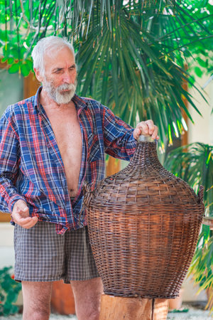 An old winemaker poses with a Large Vintage Wicker Wrapped Wine Bottle under a green palm tree. Charismatic handsome grandfather holds a bottle of Demijohn entwined with vines.の写真素材