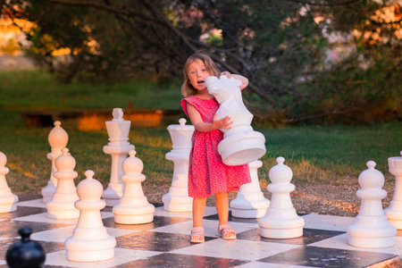 Little Caucasian girl in a pink dress playing street chess. A child plays with big chess pieces in the park on a summer evening.の写真素材