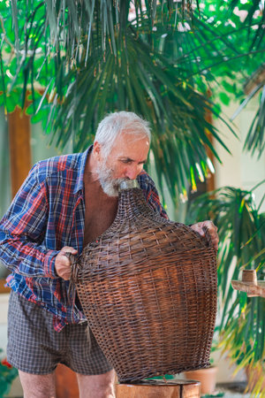 An old winemaker poses with a Large Vintage Wicker Wrapped Wine Bottle under a green palm tree. Charismatic handsome grandfather holds a bottle of Demijohn entwined with vines.の写真素材