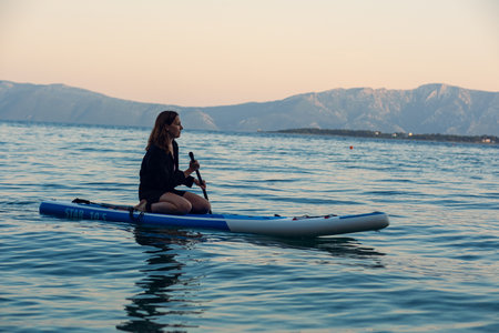 Beautiful girl swims on a sap in the sea at sunset. Beautiful calm cleaning of the sea and mountains in the background. A girl in a black shirt on a blue sap.の写真素材