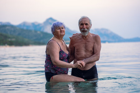 Elderly couple swims in the sea. Old bearded man and aged woman in swimsuits together stand in the tranqise water of the Adriatic sea at sunset.の写真素材